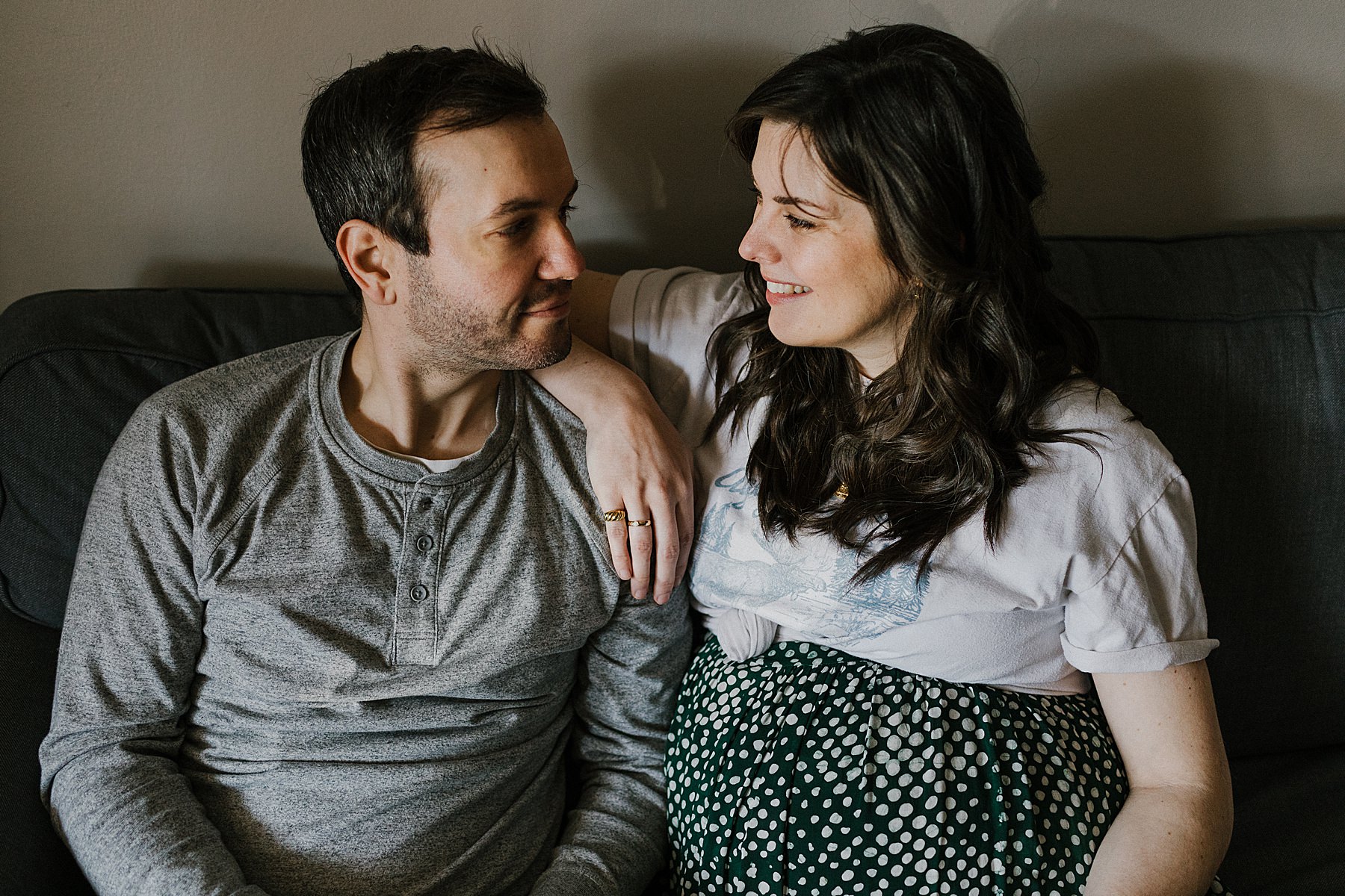 a man and a heavily pregnant woman sitting together on a sofa looking into each others eyes and smiling for pregnancy photos during maternity photography toronto bump photoshoot