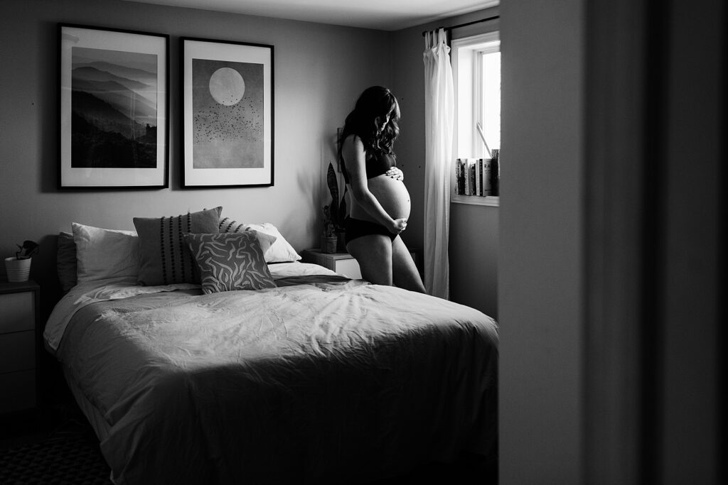 maternity photography toronto pregnancy photoshoot showing a heavily pregnant woman standing between a bed and a window holding her baby bump in mississauga GTA ontario